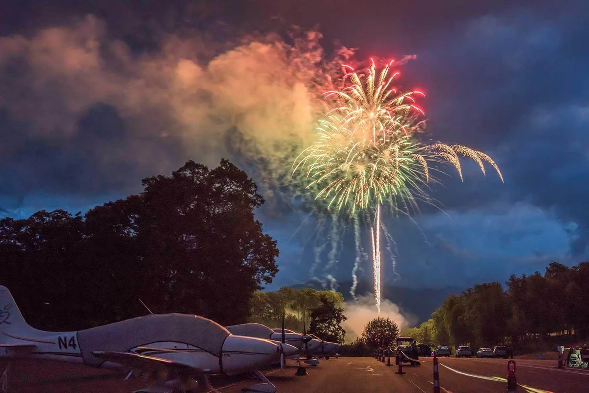 A festive exterior scene showing colorful fireworks exploding in a dark night sky, with small airplanes visible above. The mood is lively and communal, perfect for family gatherings and celebratory events.
