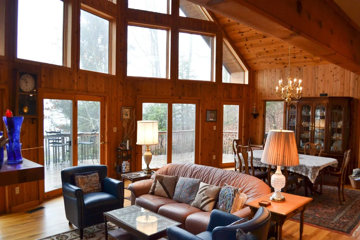 This image showcases a spacious, open living area with large windows that fill the room with natural light. The wooden walls and comfortable sofa evoke a cozy yet upscale mountain retreat atmosphere. The dining area and decorative chandelier add to the welcoming communal feel, while the windows offer glimpses of the outdoors, suggesting proximity to the surrounding landscape and amenities.