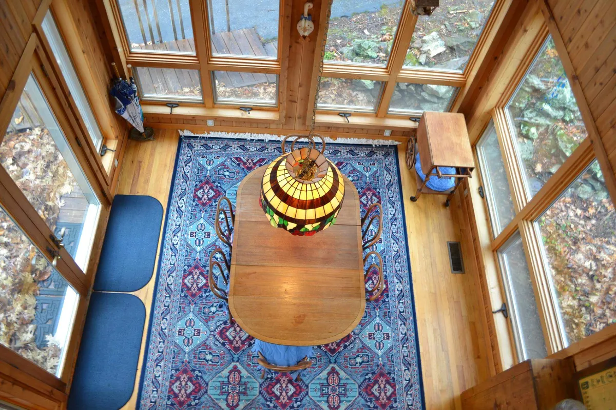 A sunlit dining area features a classic wooden table beneath a stained glass lamp, set atop an ornate rug. Large windows bring the outdoors in, hinting at the scenic surroundings and offering a gathering space for travelers and enthusiasts to connect and share their experiences.
