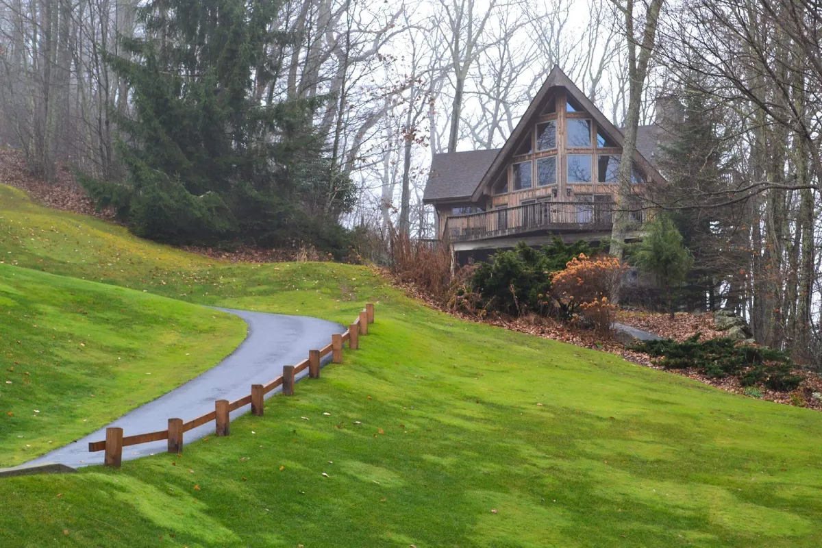The image features the exterior of a custom-built cabin nestled among lush trees, with a winding path leading up to the house. The tranquil setting emphasizes natural beauty, inviting guests to explore the garden or simply soak in the peaceful woodland surroundings. The warm, welcoming architecture hints at cozy interiors perfect for relaxation after outdoor adventures.