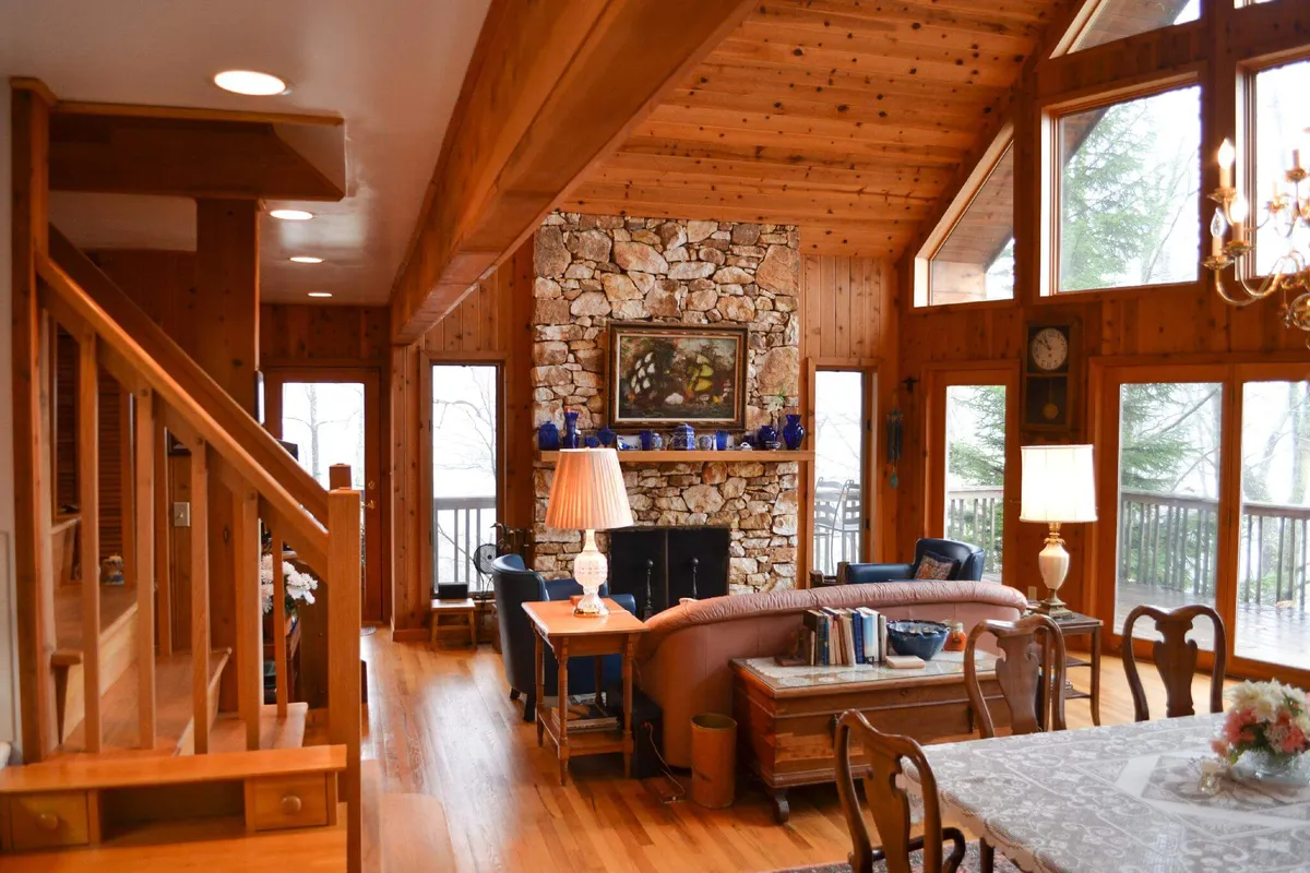 A wide-angle shot of a grand open-concept living and dining area featuring a striking stone fireplace, large windows revealing mountain views, exposed wooden beams, and a statement chandelier above a spacious dining table. The room is warmly lit, with plush seating arranged for socializing and comfortable relaxation.