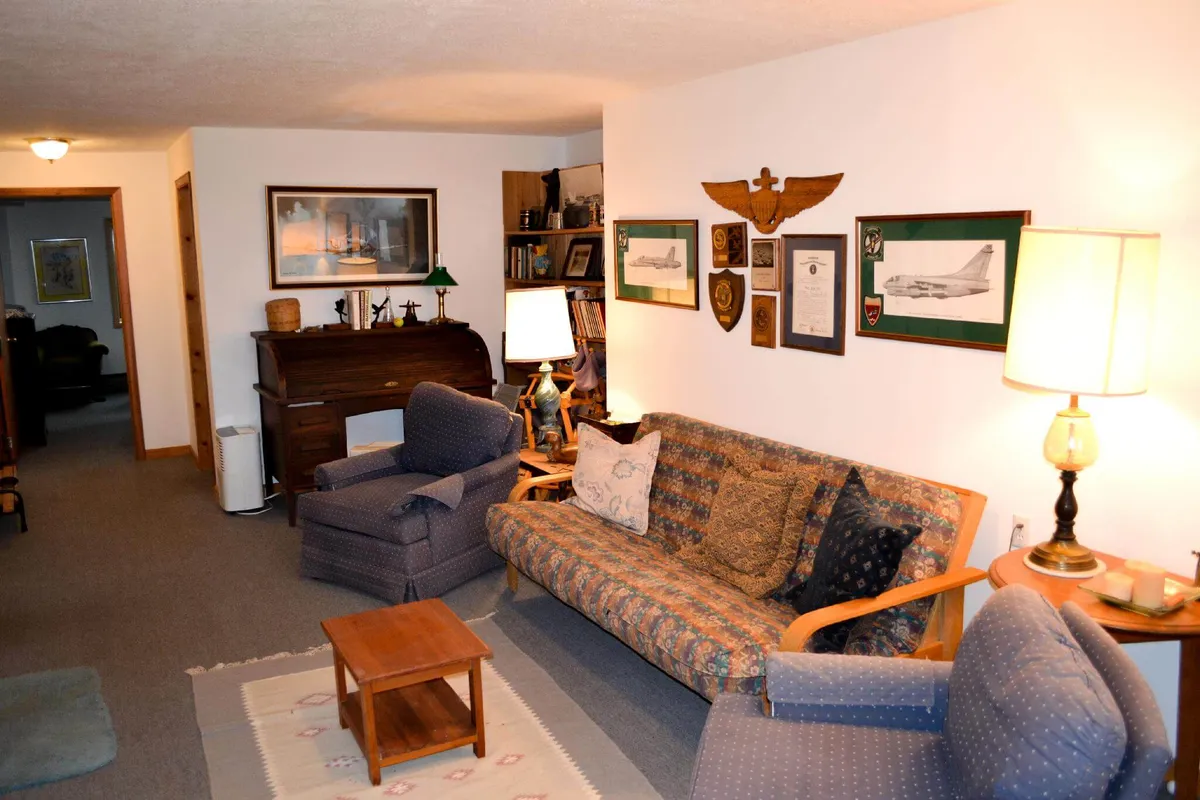 This image depicts a warm, inviting living area featuring a patterned sofa, a wooden coffee table, a softly glowing lamp, shelves filled with books, and military memorabilia adorning the wall. The space exudes a cozy, lived-in charm, perfect for unwinding with a book or enjoying meaningful conversations.
