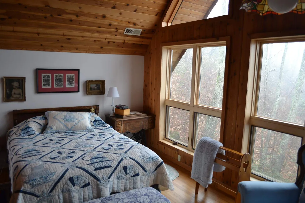 This bedroom showcases a bed with a quilt, large windows overlooking a natural view, framed artwork, and a table lamp. The scene is bright, airy, and peaceful, reflecting the text's mention of mountain breezes and restful settings.