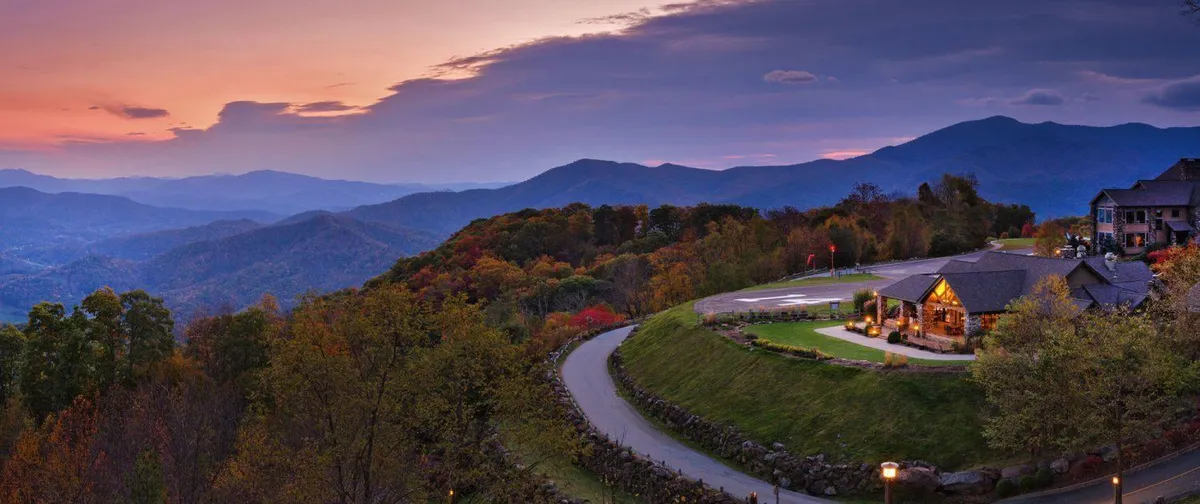 A scenic exterior shot featuring a mountain range, a lodge with a porch, and winding roads surrounded by autumn foliage. The image conveys a sense of proximity to nature, resort living, and memorable group gatherings either on the porch or nearby open spaces.