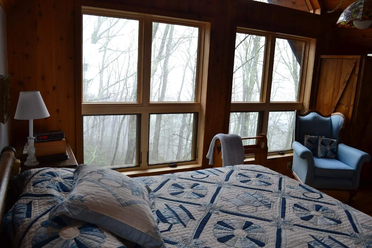 A tranquil bedroom scene with a quilted bedspread, large windows looking out onto nature, and a comfortable armchair placed for enjoying the view. The focus is on the connection between the cozy interior and the captivating natural landscape outside.