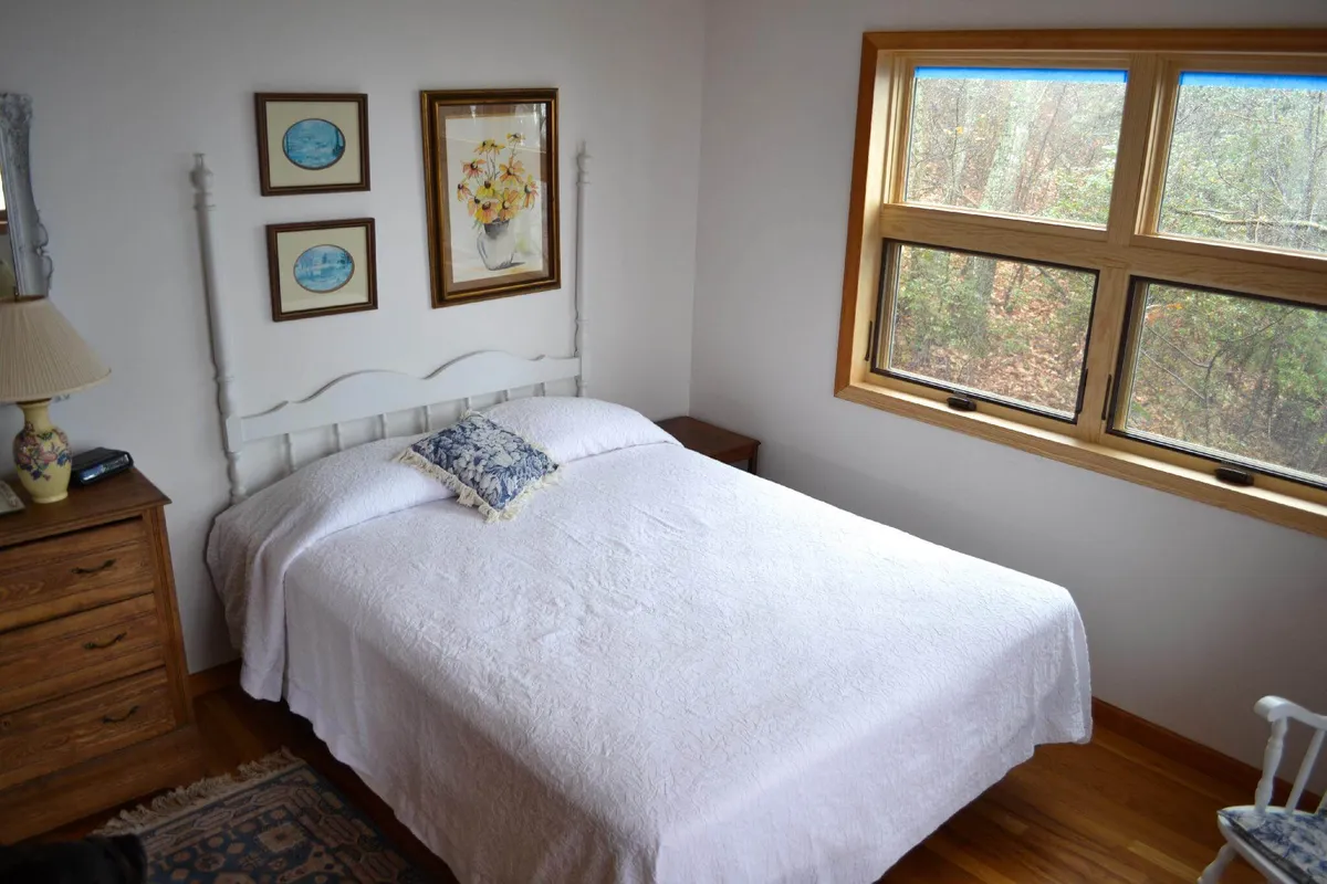 A cozy bedroom featuring a bed positioned beside a window, with soft natural light streaming in. Tasteful artwork and a lamp add to the calm, inviting atmosphere, while the window offers a glimpse of the outdoors, hinting at scenic views beyond.