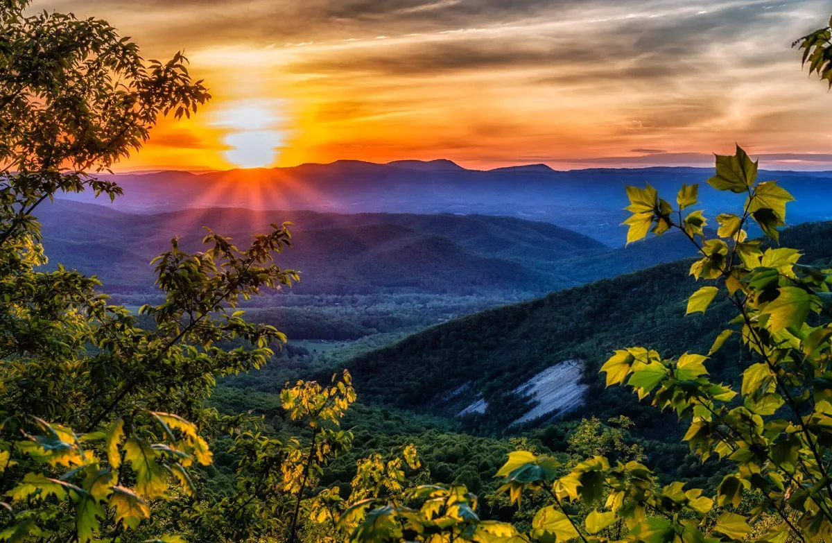 A breathtaking view captures a vivid sunset casting warm hues over a sweeping mountain range, with lush greenery visible in the foreground. The scene evokes tranquility, inviting viewers to imagine themselves enjoying the mountain air and natural beauty surrounding the resort.