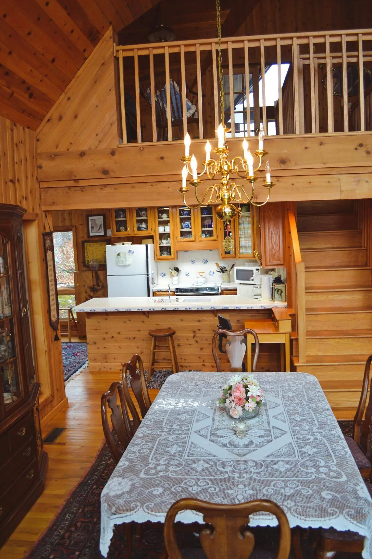 A cozy dining area featuring a wooden dining table with a lace tablecloth, an elegant chandelier overhead, a visible kitchen area in the background, and a staircase leading upward. The space exudes warmth and togetherness, highlighting the proximity between kitchen, dining, and living spaces.